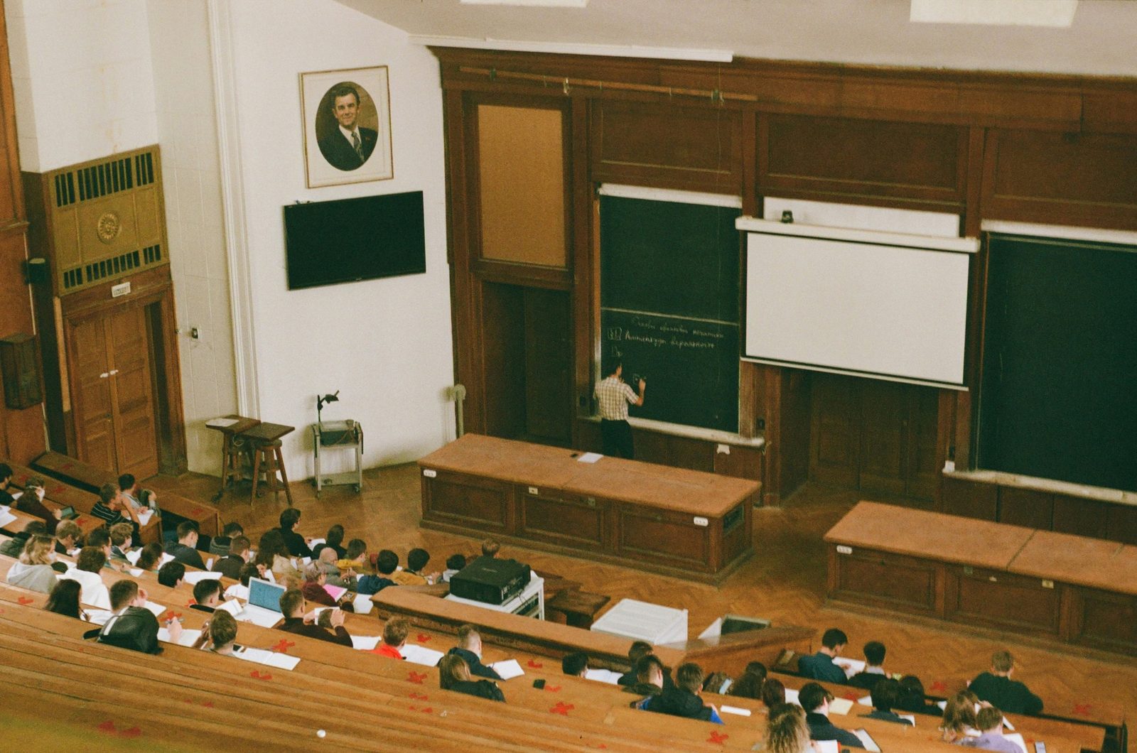 A large lecture hall seen from the upper rows: students at long wooden benches, a lecturer writing on the chalkboard.