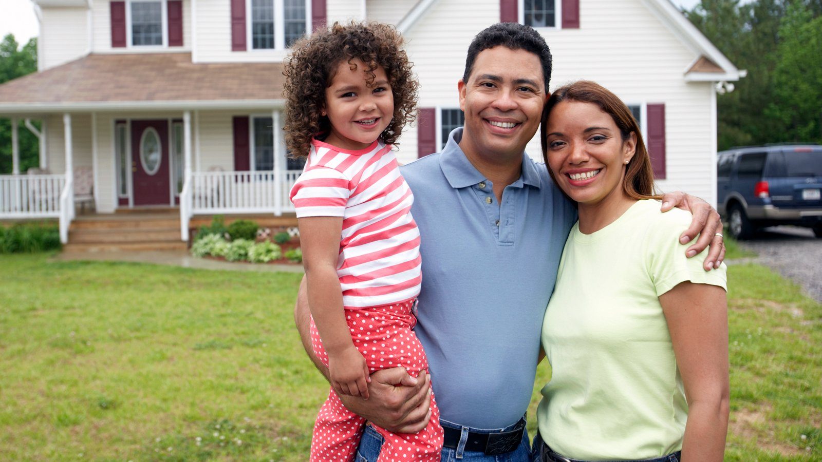 Two parents holding a young child, on the lawn in front of their house.