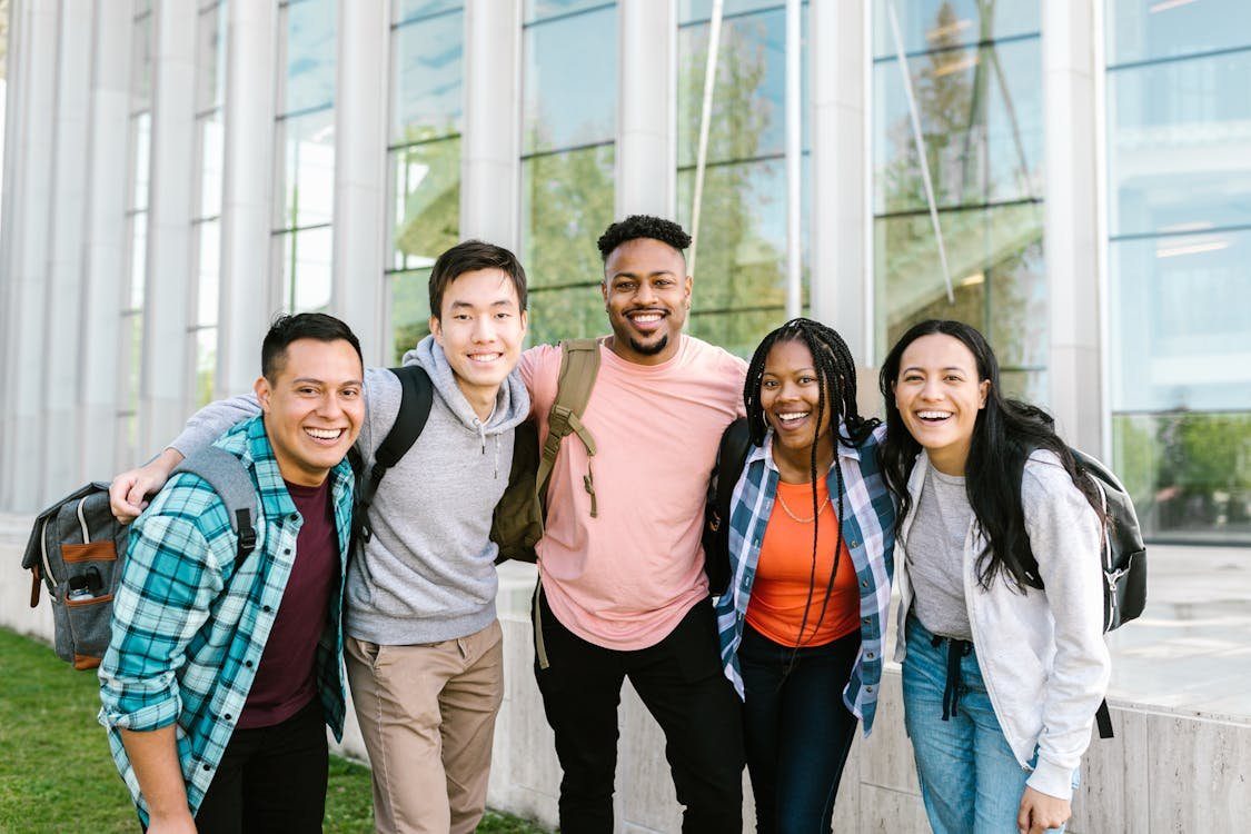 Five students with backpacks, arms around each other, outside a glass campus building.